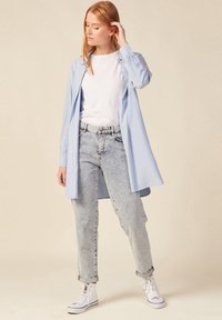 Camisa de botones azul claro sobre una camiseta blanca, combinada con jeans de tiro alto en un lavado claro y zapatillas blancas, de pie frente a un fondo neutro.