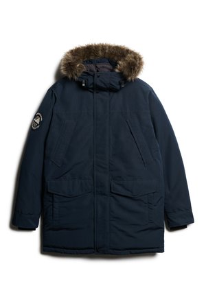 Parka azul marino con capucha forrada de piel sintética, cuatro bolsillos frontales y un logo circular en la manga izquierda. Textura suave, tejido aislante.
