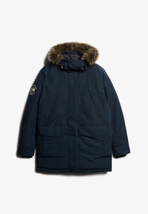 Parka blu navy con cappuccio foderato in pelliccia sintetica, quattro tasche frontali e un logo circolare sulla manica sinistra. Tessuto liscio, materiale isolante.