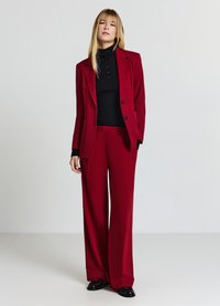Completo rosso su misura con pantaloni a gamba larga, giacca a un bottone e dolcevita nero a coste. Scarpe nere semplici completano il look.