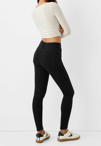 Los jeans skinny negros presentan una cinturilla alta, una textura suave y un diseño clásico de cinco bolsillos. Se combinan con zapatillas blancas y una blusa corta.