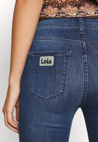 Mörkblå jeans med klassisk passform, med synliga sömmar, en bakficka med en grå etikett som säger "Lois", och subtil textur.