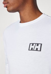 Baltas ilgomis rankovėmis marškinėliai, pagaminti iš tekstūruotos medžiagos, su juodu "HH" logotipu kairėje krūtinės pusėje. Paprasta apvalios iškirptės dizainas.