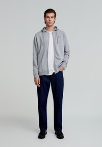 Grauer Reißverschluss-Hoodie mit Kordelzugkapuze, über einem weißen T-Shirt getragen, kombiniert mit dunklen Jeans und schwarzen Schuhen. Schlichtes Design und lässige Passform.