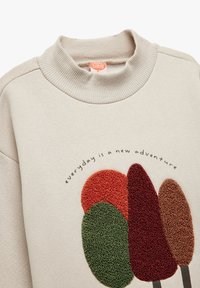 Sudadera beige con cuello alta acanalado que presenta formas de árboles bordadas en rojo, verde y marrón con el texto "cada día es una nueva aventura."