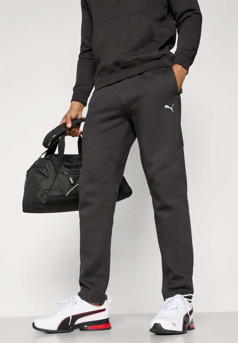 Joggers atléticos negros con un ajuste elegante, con un pequeño logotipo blanco, combinados con zapatillas negras y blancas y una bolsa de deporte negra.