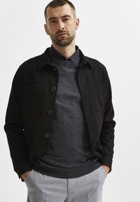 Black button-up jacket with a textured finish, layered over a dark gray sweater. Gray trousers with a smooth, tailored look.