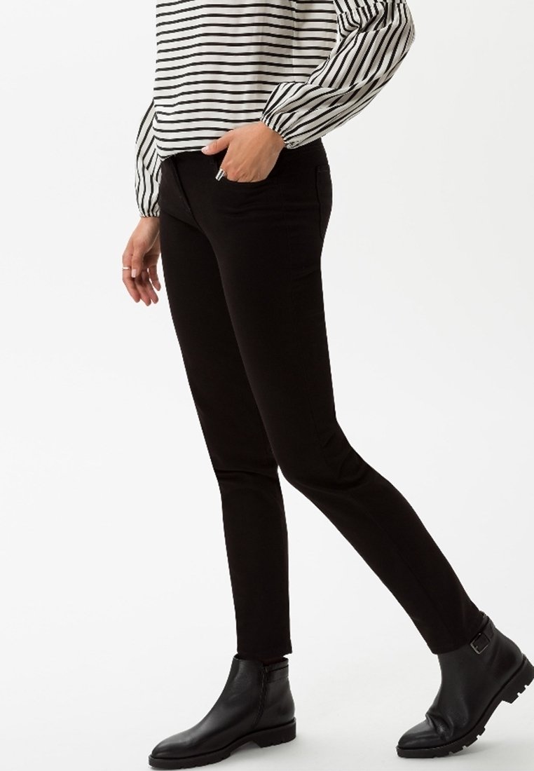Personne portant un pantalon slim noir, des bottines noires et une chemise blanche à manches longues avec des rayures horizontales noires.