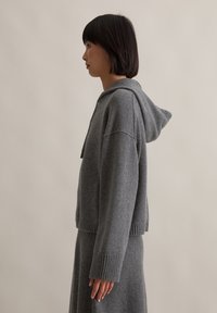 Grauer gestrickter Hoodie mit geripptem Saum und Bündchen, ausgestattet mit einer verstellbaren Kapuze. Weiche Textur, cropped Design, gezeigt mit passendem grauem Rock.