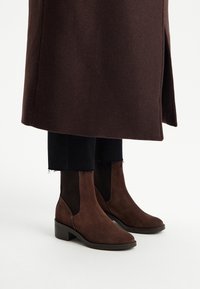 Brązowe zamszowe botki za kostkę z elastycznymi panelami bocznymi, niskim, warstwowym obcasem i gładką fakturą, w zestawieniu z czarnymi spodniami i brązowym płaszczem.