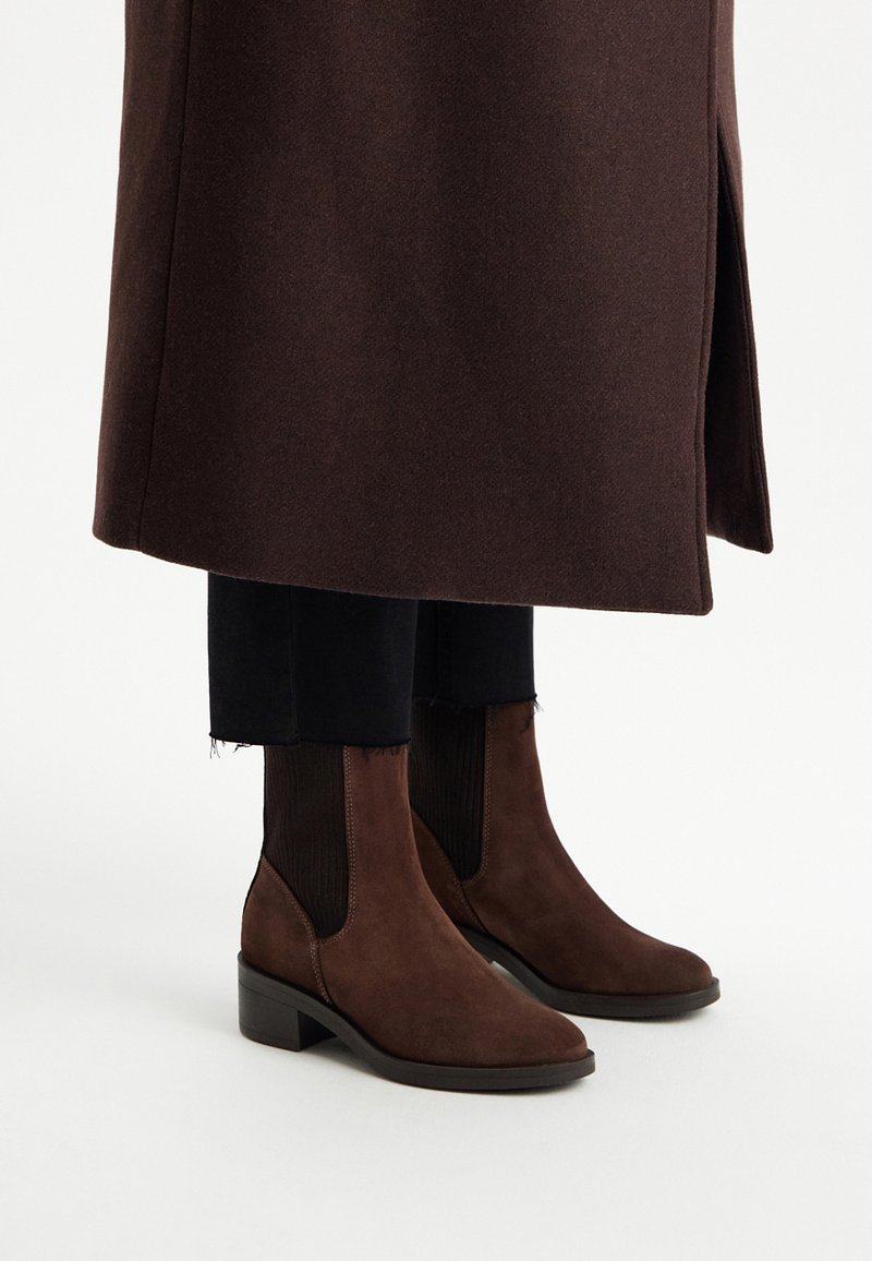 Brązowe zamszowe botki za kostkę z elastycznymi panelami bocznymi, niskim, warstwowym obcasem i gładką fakturą, w zestawieniu z czarnymi spodniami i brązowym płaszczem.