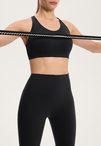 Czarna sportowa biustonosz i legginsy o wysokim stanie wykonane z gładkiego, elastycznego materiału. Modelka trzyma taśmę oporową z teksturowaną powierzchnią.