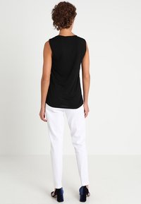 Top noir sans manches avec une coupe ample et un dos drapé, associé à un pantalon blanc bien taillé et des chaussures à talons carrés bleues.