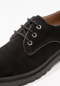 Scarpa in suede nera con una texture liscia, punta arrotondata e cuciture chiare a contrasto. Presenta tre occhielli e una suola in gomma resistente.