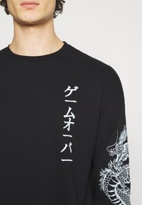 Maglia nera a maniche lunghe con testo giapponese bianco sulla parte anteriore e grafica di un drago sulla manica destra. Tessuto di cotone, vestibilità classica.