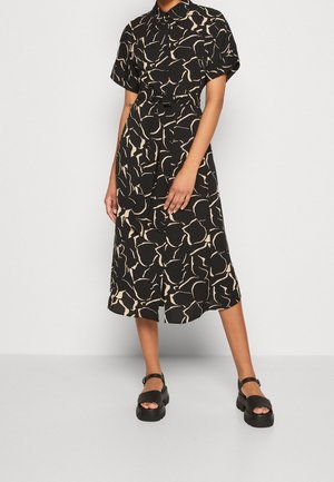 Kobieta prezentująca czarną midi sukienkę z abstrakcyjnymi beżowymi wzorami, krótkimi rękawami, paskiem w talii oraz czarnymi sandałami na masywnych paskach.