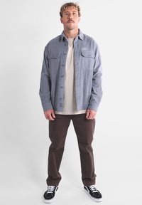Camisa de tejido gris con dos bolsillos en el pecho, superpuesta a una camiseta ligera beige, combinada con pantalones marrones y zapatillas negras. El modelo está de pie.