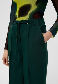 Green tailored trousers with a drawstring waist, side pockets, and soft-textured fabric, featuring a structured silhouette and pleated details.