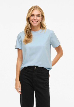 Giovane donna con capelli biondi lunghi che indossa una maglietta a maniche corte azzurro chiaro e jeans neri a vita alta, sorridente con una mano dietro la schiena.