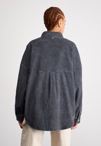 Šedá kordurová košeľa s voľným strihom, ktorá má zadočku, dlhé rukávy a gombíkové manžety. Tkanina má rebrovanú textúru.