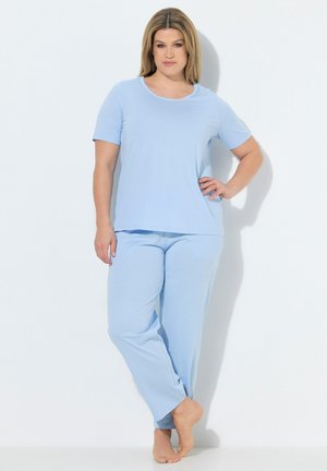 Femme debout pieds nus sur fond blanc, portant un haut bleu clair à manches courtes et un pantalon ample assorti avec un motif subtil à pois.