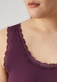 Top a canotta a costine di un profondo viola con pizzo lungo il collo e le spalle, caratterizzato da una texture liscia e un design aderente.