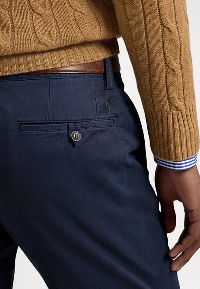Granatowe spodnie z tylną kieszenią zapinaną na guzik. Noszone z beżowym swetrem w warkoczowy splot i mankietami koszuli w paski.