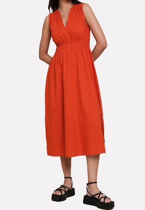 Ärmelloses, oranges Midikleid mit V-Ausschnitt und geraffter Taille, getragen mit schwarzen Plateausandalen an einer stehenden Person.