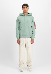 Mentazöld kapucnis pulóver kenguruzsebbel, állítható zsinórral ellátott kapucnival és logóval. Bézs cargo nadrággal és világos sportcipővel párosítva.