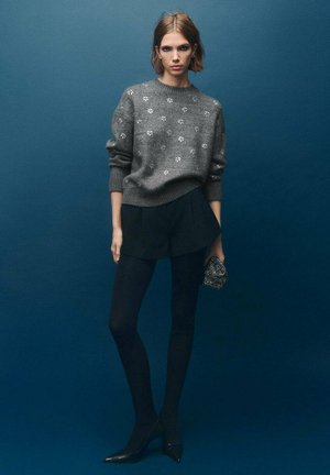 Suéter gris con patrones florales, falda negra, medias negras y tacones de punta. Sosteniendo un pequeño bolso de cuentas. Fondo oscuro y sólido.