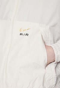 White lightweight jacket with a zip front, ribbed cuffs, featuring a gold Nike swoosh and black "AIR" logo on the chest. Soft texture.