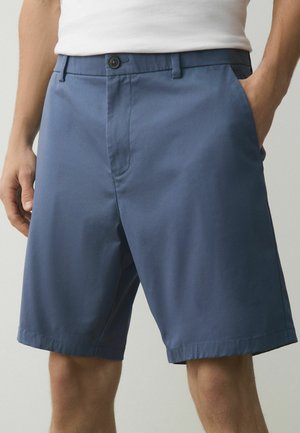 Középhosszú kék rövidnadrágot és fehér inget viselő férfi, egyik kezét a zsebében tartva, semleges háttér előtt áll.