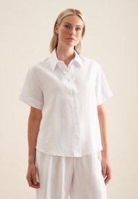 Camicetta bianca in lino a maniche corte con colletto, chiusura frontale a bottoni e una vestibilità rilassata, caratterizzata da una sottile texture e linee pulite.