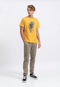 Geel katoenen t-shirt met een grafische afbeelding van een hand, gecombineerd met beige cargobroek en zwarte sneakers, tegen een witte achtergrond.