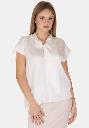 Giovane donna con capelli biondi lisci, che indossa una blusa bianca a maniche corte con dettaglio a laccio sul davanti e una gonna chiara, su uno sfondo neutro.