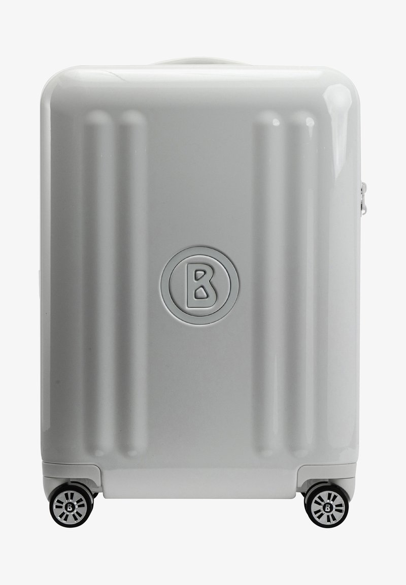 Valise rigide de couleur gris clair avec des rainures horizontales, un logo circulaire et quatre roues noires pour la mobilité. Texture lisse et fermeture éclair.