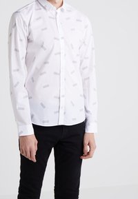 Weißes Hemd mit Button-Up-Verschluss, das ein dezentes geometrisches Muster in Grau aufweist, lange Ärmel und einen normalen Kragen hat. Kombiniert mit schwarzen Hosen.