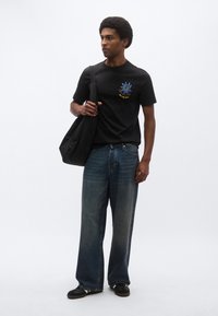 Μαύρο μπλουζάκι με μπλε και κίτρινο γραφικό σχέδιο, συνδυασμένο με φαρδιά ξεθωριασμένα μπλε τζιν και μαύρα αθλητικά παπούτσια. Κρατάει μια μαύρη τσάντα ώμου.