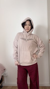 Γυναίκα με μπεζ φούτερ με κουκούλα που έχει μαύρο φυλετικό σχέδιο καρδιάς, κόκκινο και μαύρο καρό παντελόνι, στέκεται με το ένα χέρι στη μέση μπροστά από απλό τοίχο.