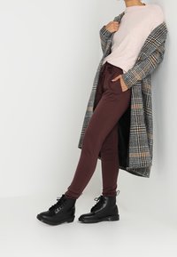 Langer kariert Mantel in Grau und Schwarz mit orangenen Akzenten, hellrosa Pullover, bordeauxfarbene Hose und schwarze Schnürstiefel.