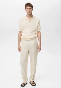 Korte mouw, crèmekleurig gebreid polo shirt met een drie-knopen sluiting, gecombineerd met lichtbeige, op maat gemaakte broeken en zwarte slippers.