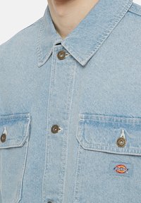 Ljusblå denimjacka med krage, framknäppning med knappar, två bröstfickor och en röd och gul Dickies-logopatch.