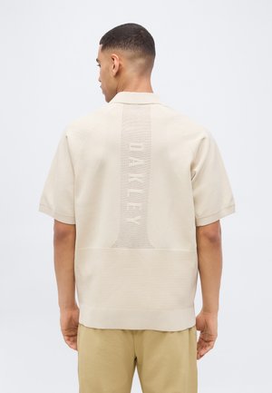 Vyras trumpais plaukais, dėvintis smėlio spalvos trumpomis rankovėmis megztą marškinėlį su tekstūruotu vertikaliu "OAKLEY" tekstu ant nugaros ir derančias smėlio spalvos kelnes.