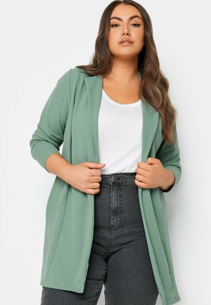 Giacca lunga e aperta davanti in morbido tessuto verde chiaro con una vestibilità rilassata. Presenta maniche lunghe e spacchi sui lati. Indossata sopra un top bianco e jeans scuri.