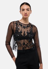 Haut transparent noir à manches longues, présentant des perles noires délicates et des broderies florales, superposé sur une couche inférieure noire unie.