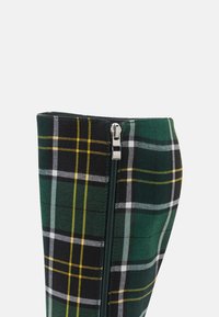 Bottine à carreaux verts et noirs avec des accents jaunes, dotée d'une fermeture éclair sur le côté et d'une texture lisse au toucher.