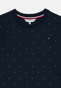 Sweatshirt azul-marinho com um decote redondo, salpicado de pequenos studs prateados e uma etiqueta em vermelho, branco e azul perto do colarinho.