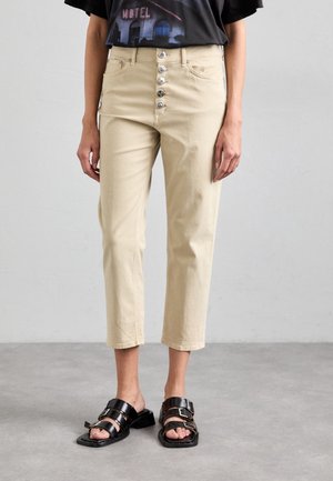 Beige Cropped-Hosen mit vier sichtbaren vorderen Knöpfen, getragen mit schwarzen offenen Sandalen mit zwei breiten Riemen und Schnallen.