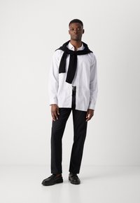Cămașă albă cu dungi, pulover negru legat în jurul umerilor, pantaloni negri și pantofi negri. Design simplu, modern, cu accente minime.