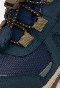 Gros plan sur une basket en tissu bleu et en daim avec des boucles de lacets beiges et des lacets bruns à motifs sur une semelle claire.